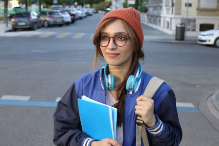 Photo for Student woman wearing beanie hat and eyeglasses holding notebooks walking outdoors - Royalty Free Image