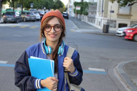 Téléchargez les photos : Étudiante portant un bonnet et des lunettes tenant des cahiers marchant à l'extérieur - en image libre de droit
