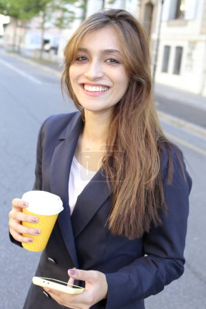 Photo for Businesswoman in suit holding smartphone and coffee outdoors - Royalty Free Image