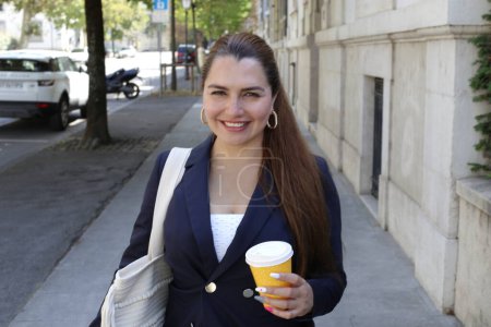 Téléchargez les photos : Portrait de belle jeune femme d'affaires confiante avec tasse de café en papier sur la rue de la ville - en image libre de droit