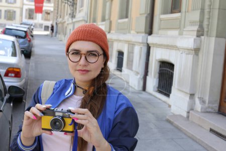 Foto de Retrato de mujer hermosa joven en ropa hipster con estilo con auriculares vintage y cámara de cine en la calle de la ciudad - Imagen libre de derechos