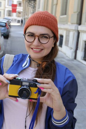 Foto de Retrato de mujer hermosa joven en ropa hipster con estilo con auriculares vintage y cámara de cine en la calle de la ciudad - Imagen libre de derechos