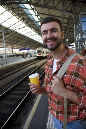 Téléchargez les photos : Portrait de beau jeune homme heureux attendant le train à la gare tout en voyageant avec sac à dos - en image libre de droit