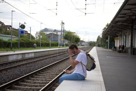 Foto de Retrato de joven feliz guapo esperando tren en la estación mientras viaja con mochila - Imagen libre de derechos