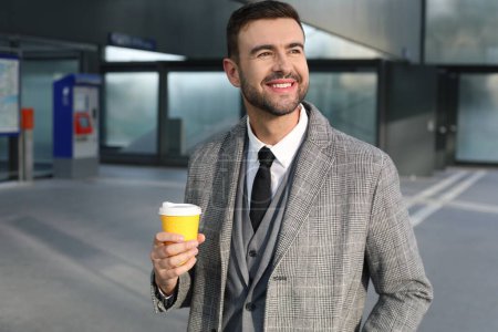 Téléchargez les photos : Portrait de beau jeune homme en costume gris avec tasse de café en papier sur la rue de la ville - en image libre de droit
