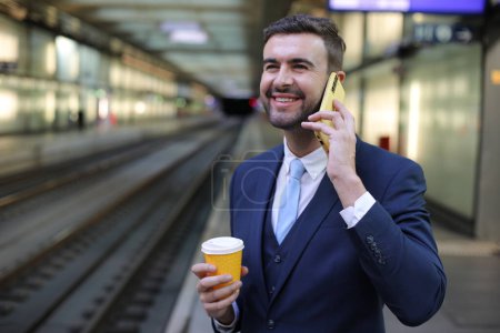 Téléchargez les photos : Portrait de beau jeune homme d'affaires en costume avec tasse en papier de café et smartphone sur la gare - en image libre de droit