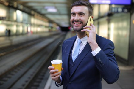 Foto de Retrato de joven hombre de negocios guapo en traje con taza de papel de café y teléfono inteligente en la estación de tren - Imagen libre de derechos