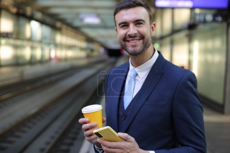Photo for Portrait of handsome young businessman in suit with paper cup of coffee and smartphone on train station - Royalty Free Image