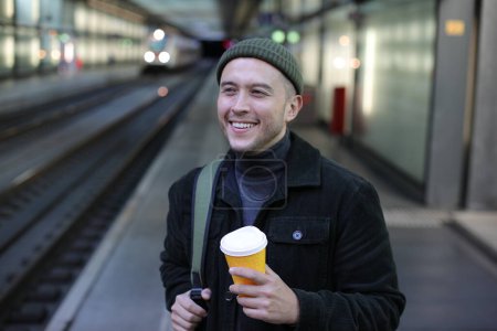 Foto de Retrato de joven guapo en ropa elegante con taza de papel de café en la estación de tren - Imagen libre de derechos