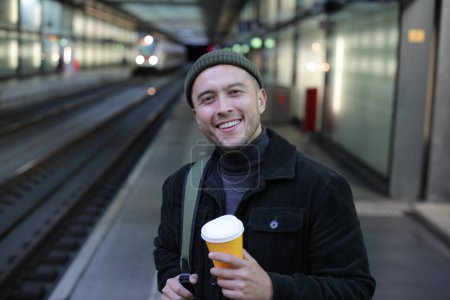 Foto de Retrato de joven guapo en ropa elegante con taza de papel de café en la estación de tren - Imagen libre de derechos