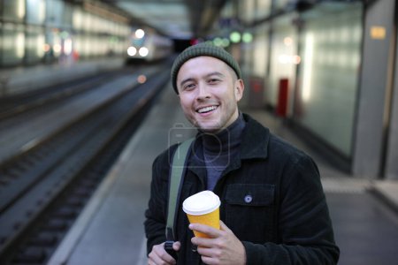 Téléchargez les photos : Portrait de beau jeune homme en vêtements élégants avec tasse de café en papier sur la gare - en image libre de droit