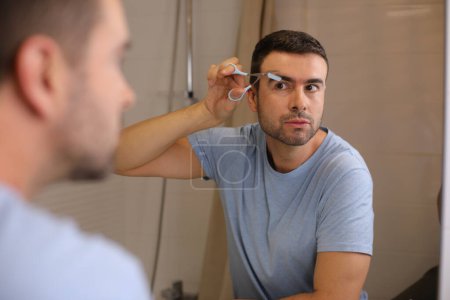 Photo for Portrait of handsome young man shaving his eyebrows in front of mirror in bathroom - Royalty Free Image