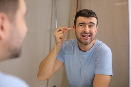 Téléchargez les photos : Portrait de beau jeune homme nettoyant les oreilles devant le miroir dans la salle de bain - en image libre de droit
