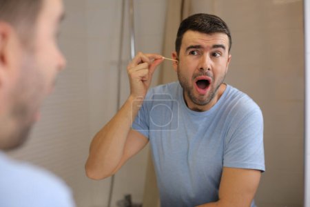 Téléchargez les photos : Portrait de beau jeune homme nettoyant les oreilles devant le miroir dans la salle de bain - en image libre de droit