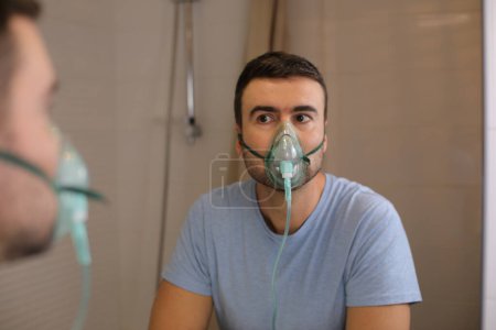 Téléchargez les photos : Portrait de beau jeune homme avec masque à oxygène devant le miroir dans la salle de bain - en image libre de droit