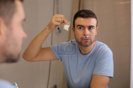 Téléchargez les photos : Portrait d'un beau jeune homme tenant un préservatif devant un miroir dans la salle de bain - en image libre de droit