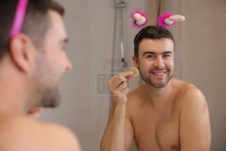 Téléchargez les photos : Portrait d'un beau jeune homme tenant un préservatif devant un miroir dans la salle de bain - en image libre de droit