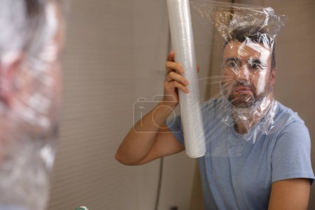 Foto de Retrato de un joven guapo cubriéndose la cabeza con cinta de plástico delante del espejo en el baño - Imagen libre de derechos