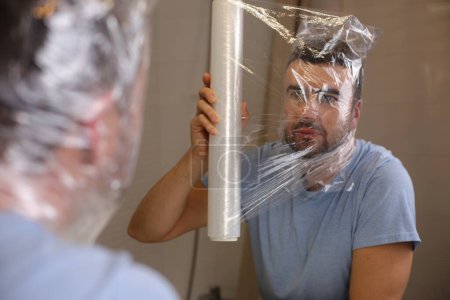 Foto de Retrato de un joven guapo cubriéndose la cabeza con cinta de plástico delante del espejo en el baño - Imagen libre de derechos