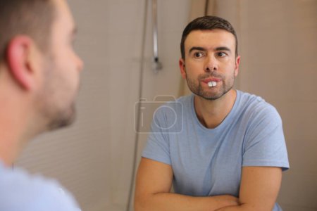 Téléchargez les photos : Portrait de beau jeune homme avec de grandes dents devant le miroir dans la salle de bain - en image libre de droit