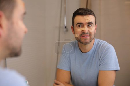 Téléchargez les photos : Portrait de beau jeune homme avec bande de bandage sur le visage devant le miroir dans la salle de bain - en image libre de droit