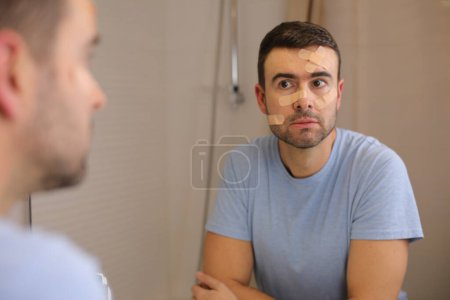 Téléchargez les photos : Portrait de beau jeune homme avec bande de bandage sur le visage devant le miroir dans la salle de bain - en image libre de droit