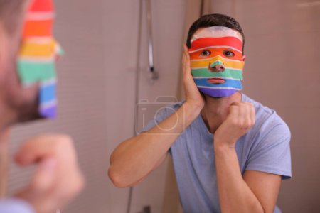 Foto de Retrato de joven guapo con máscara facial en colores de la bandera lgbtqa delante del espejo en el baño - Imagen libre de derechos