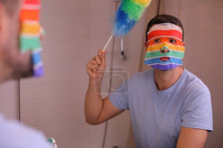 Téléchargez les photos : Portrait de beau jeune homme avec masque de soins du visage aux couleurs du drapeau lgbtqa devant le miroir dans la salle de bain - en image libre de droit