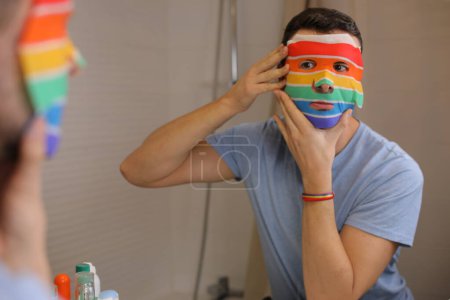 Photo for Portrait of handsome young man with facecare mask in colors of lgbtqa flag in front of mirror in bathroom - Royalty Free Image