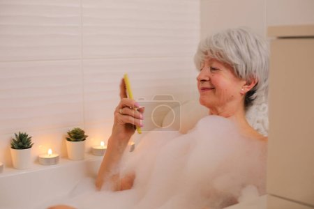 Téléchargez les photos : Portrait de femme âgée heureuse avec smartphone dans la baignoire avec bulles de savon - en image libre de droit