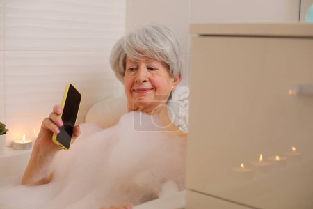 Téléchargez les photos : Portrait de femme âgée heureuse avec smartphone dans la baignoire avec bulles de savon - en image libre de droit