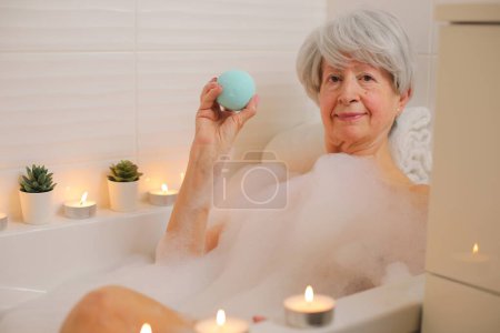 Foto de Retrato de mujer mayor feliz relajarse en la bañera con burbujas de jabón y la celebración de la bomba de baño - Imagen libre de derechos