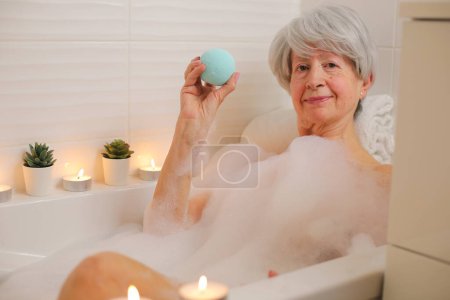 Téléchargez les photos : Portrait de femme âgée heureuse se détendre dans la baignoire avec des bulles de savon et tenant la bombe de bain - en image libre de droit