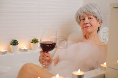 Téléchargez les photos : Portrait de femme âgée heureuse avec verre de vin rouge dans la baignoire avec bulles de savon - en image libre de droit