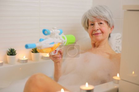 Téléchargez les photos : Portrait de femme âgée heureuse avec pistolet à eau dans la baignoire avec bulles de savon - en image libre de droit
