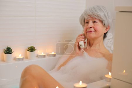 Téléchargez les photos : Portrait de femme âgée heureuse parlant par téléphone dans la baignoire avec des bulles de savon - en image libre de droit