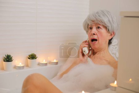 Téléchargez les photos : Portrait de femme âgée heureuse parlant par téléphone dans la baignoire avec des bulles de savon - en image libre de droit