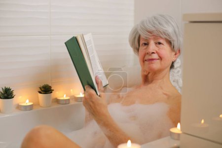 Téléchargez les photos : Portrait de femme âgée heureuse relaxant et livre de lecture dans la baignoire avec bulles de savon - en image libre de droit