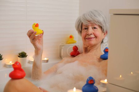 Téléchargez les photos : Portrait de femme âgée heureuse se détendre dans la baignoire avec des bulles de savon et des canards en caoutchouc - en image libre de droit