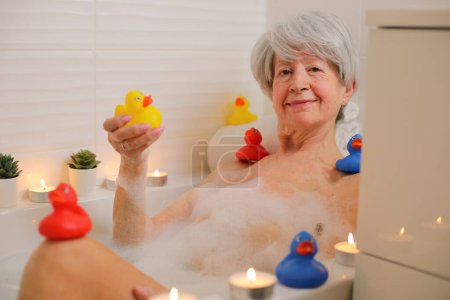 Téléchargez les photos : Portrait de femme âgée heureuse se détendre dans la baignoire avec des bulles de savon et des canards en caoutchouc - en image libre de droit