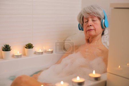 Téléchargez les photos : Portrait de femme âgée heureuse se détendre dans la baignoire avec des bulles de savon et écouter de la musique avec des écouteurs - en image libre de droit