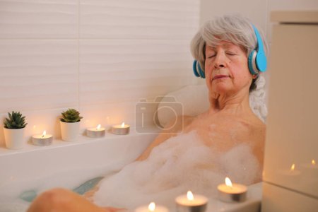 Téléchargez les photos : Portrait de femme âgée heureuse se détendre dans la baignoire avec des bulles de savon et écouter de la musique avec des écouteurs - en image libre de droit