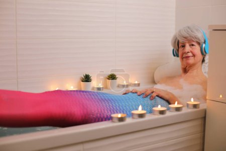 Téléchargez les photos : Portrait de femme âgée heureuse avec queue de sirène relaxant dans la baignoire avec bulles de savon et écouter de la musique avec écouteurs - en image libre de droit