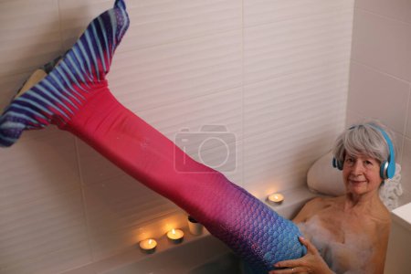 Foto de Retrato de mujer mayor feliz con cola de sirena relajándose en la bañera con burbujas de jabón y escuchando música con auriculares - Imagen libre de derechos