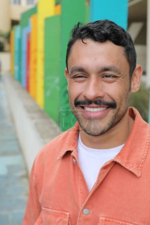 Téléchargez les photos : Portrait d'homme hispanique souriant heureux avec moustache sur la rue de la ville en regardant la caméra en face de fond coloré - en image libre de droit