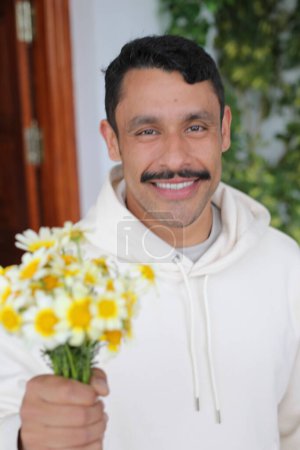 Foto de Retrato de hombre hispano guapo con bigote en sudadera blanca sonriendo y dando ramo de manzanillas a cámara - Imagen libre de derechos