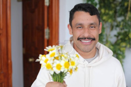 Foto de Retrato de hombre hispano guapo con bigote en sudadera blanca sonriendo y dando ramo de manzanillas a cámara - Imagen libre de derechos