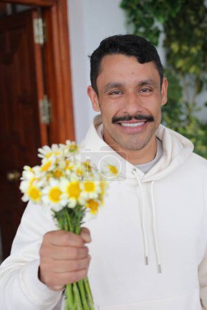 Foto de Retrato de feliz hombre hispano con bigote con capucha blanca sonriendo y dando ramo de manzanillas a cámara - Imagen libre de derechos