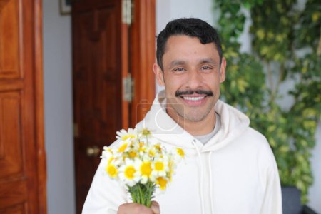 Foto de Retrato de feliz hombre hispano con bigote con capucha blanca sonriendo y dando ramo de manzanillas a cámara - Imagen libre de derechos