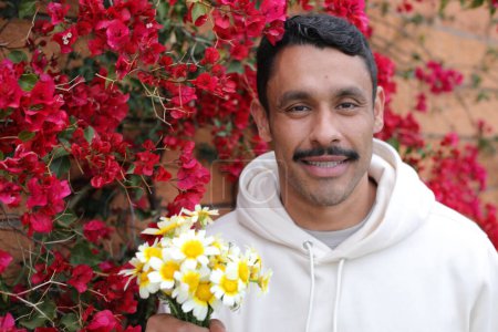 Téléchargez les photos : Portrait d'un bel homme hispanique avec moustache en sweat à capuche blanc tenant un bouquet de camomilles - en image libre de droit